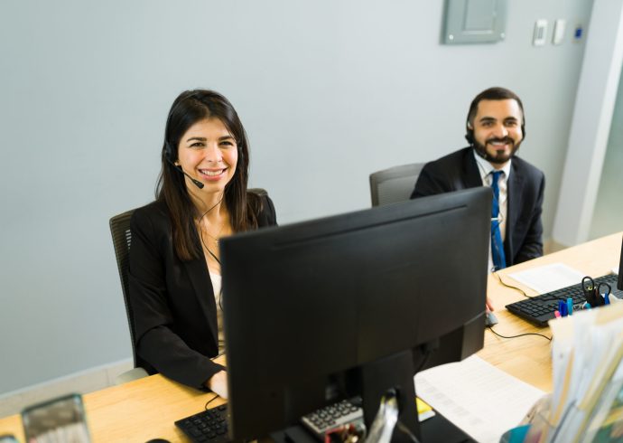 Happy co-workers smiling and making eye contact while sitting at their desk in a call center. Executive colleagues waiting to receive calls from customers for tech support